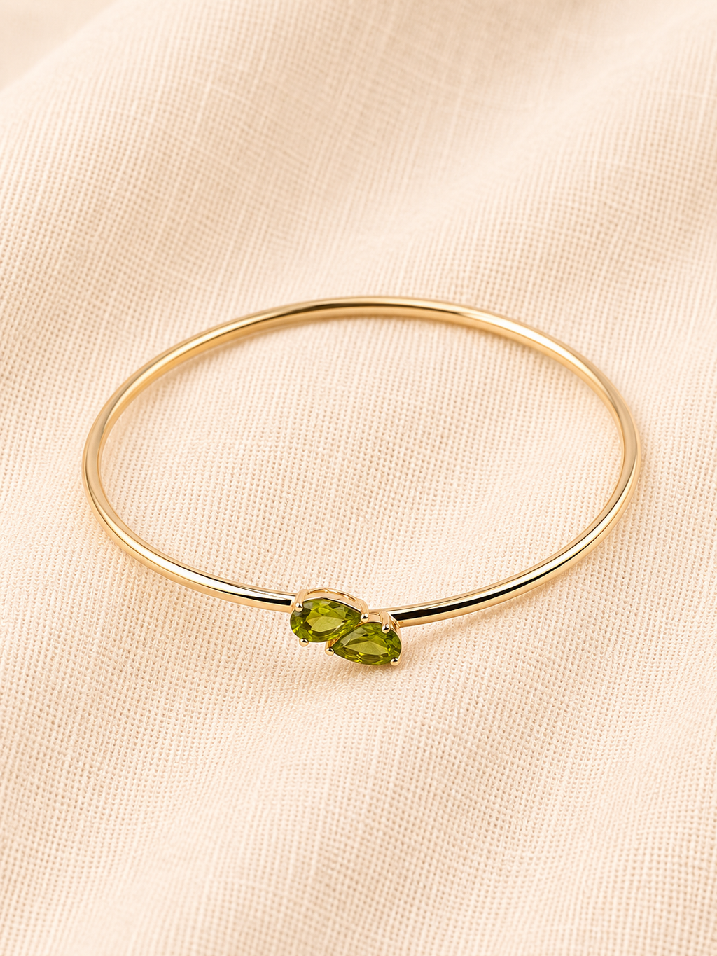 A solid 10k yellow gold bangle bracelet with two natural green peridot gemstone accent in teardrop shape by Ophelisse, displayed on a textured beige fabric background, showing flat top angle view.