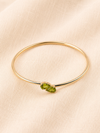 A solid 10k yellow gold bangle bracelet with two natural green peridot gemstone accent in teardrop shape by Ophelisse, displayed on a textured beige fabric background, showing flat top angle view.