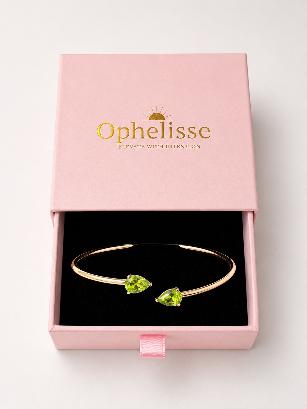 A solid 10k yellow gold open cuff bracelet with two natural green pear-cut peridot gemstones, presented in Ophelisse pink jewelry box printed in gold, showing the top angle view of the bangle.