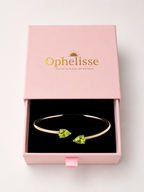 A solid 10k yellow gold open cuff bracelet with two natural green pear-cut peridot gemstones, presented in Ophelisse pink jewelry box printed in gold, showing the top angle view of the bangle.