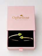 A solid 10k gold bangle bracelet with two natural green teardrop peridot gemstones, presented in an open pink Ophelisse gift box with a black interior, showing the top side angle view.