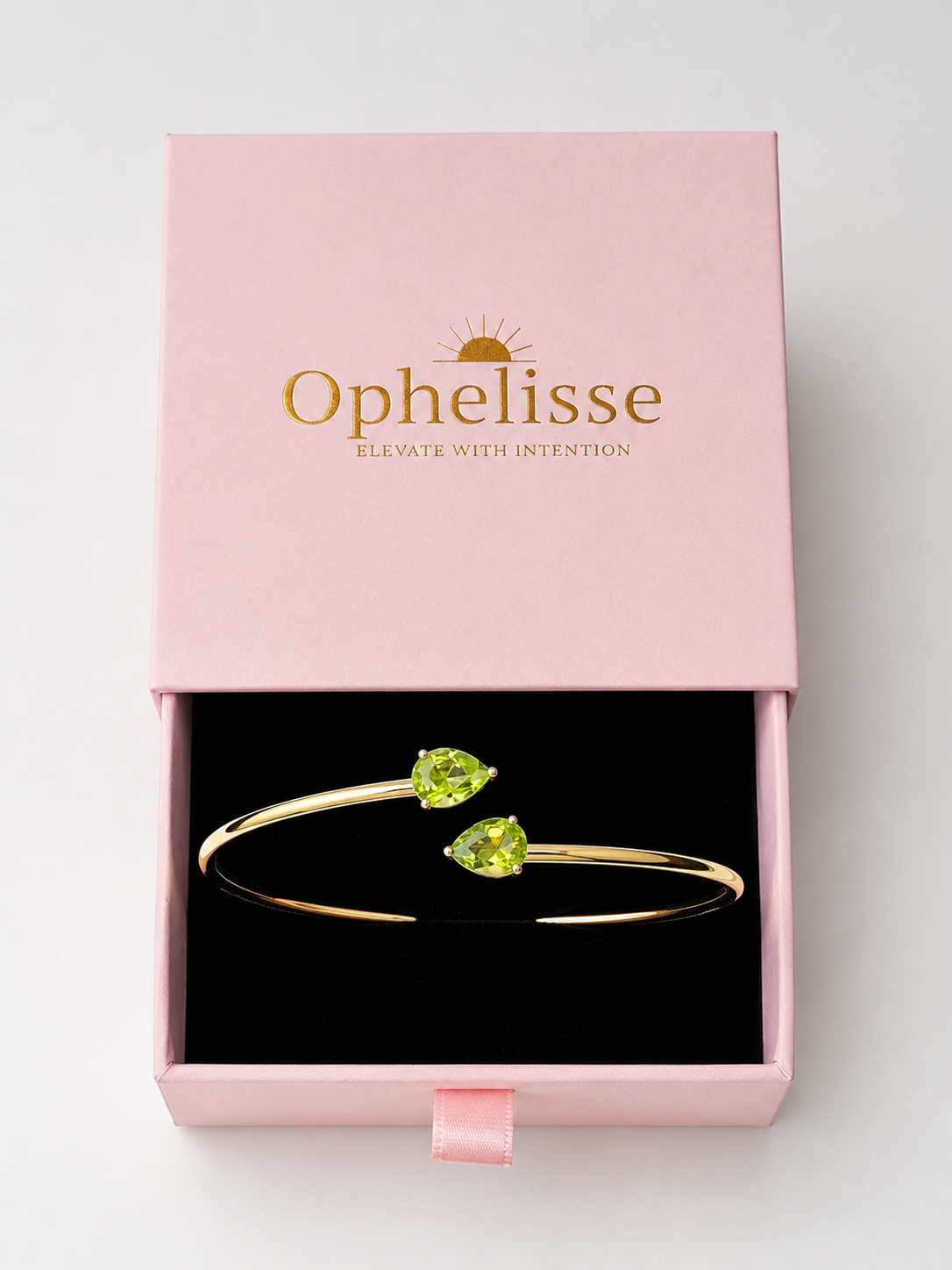 A solid 10k gold bangle bracelet with two natural green teardrop peridot gemstones, presented in an open pink Ophelisse gift box with a black interior, showing the top side angle view.