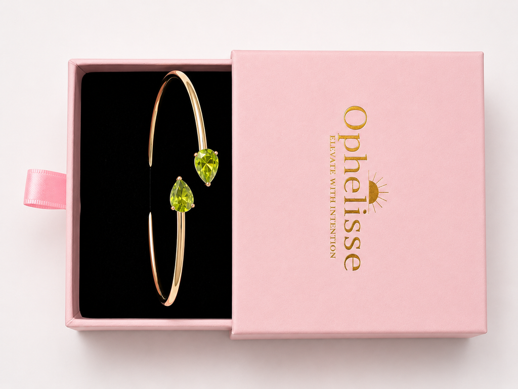 Top view of a solid 10k yellow gold bangle bracelet with two natural green heart-shaped peridot gemstones, presented in an open pink gift box with the lid showing the brand name Ophelisse.