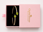 Top view of a solid 10k yellow gold bangle bracelet with two natural green heart-shaped peridot gemstones, presented in an open pink gift box with the lid showing the brand name Ophelisse.