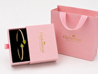 A solid 10k gold bangle bracelet with two green pear-shaped natural peridot gemstones displayed in an open pink Ophelisse gift box, next to a pink Ophelisse shopping bag on a white background - an Ophelisse luxurious unpackaging experience.