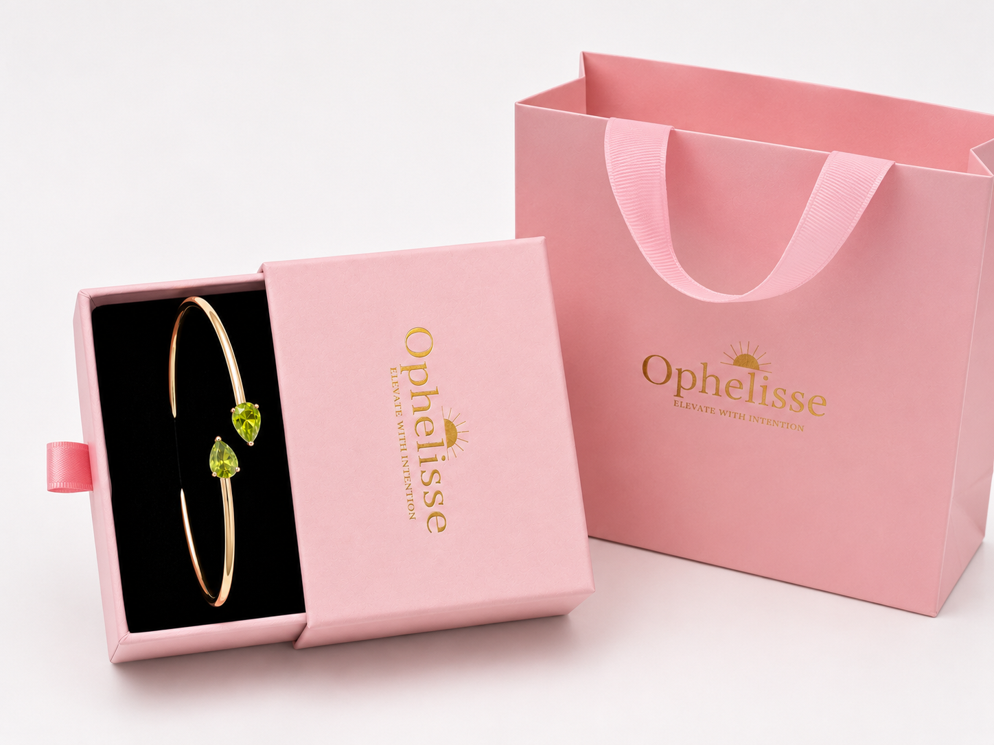 A solid 10k gold bangle bracelet with two green pear-shaped natural peridot gemstones displayed in an open pink Ophelisse gift box, next to a pink Ophelisse shopping bag on a white background - an Ophelisse luxurious unpackaging experience.