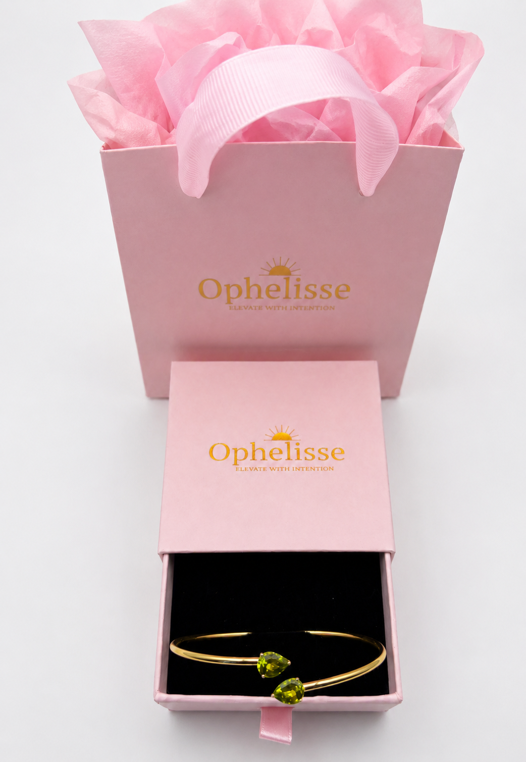A solid 10k yellow gold bangle bracelet with two natural green peridot gemstone accents displayed in an open pink Ophelisse jewelry box, with the matching pink branded gift bag and tissue paper behind it on a white background.