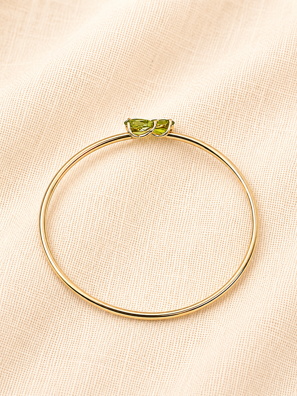 A solid 10k yellow gold bangle bracelet with two natural green peridot gemstone set at the top, displayed on a textured beige fabric background.