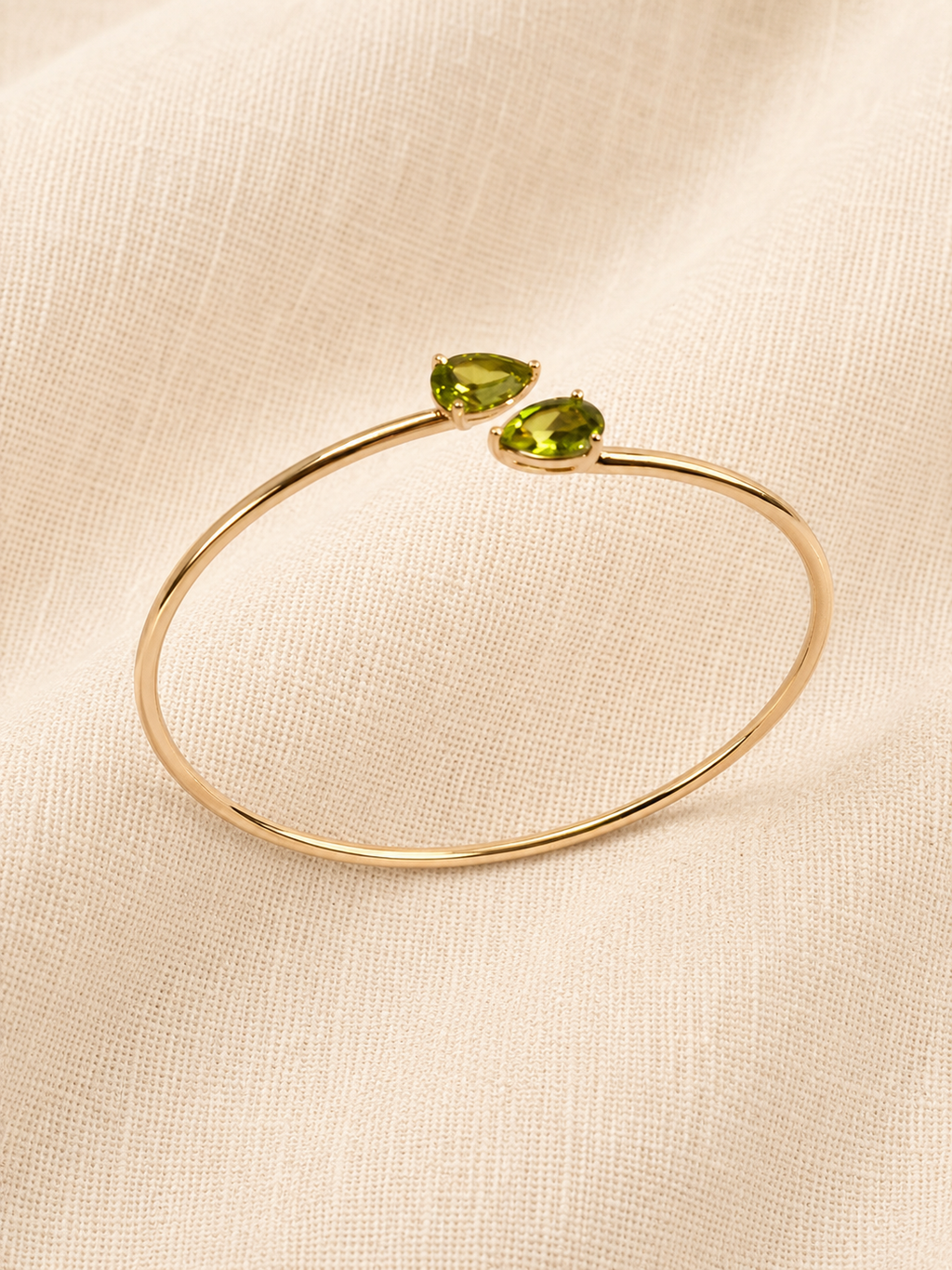 A solid 10k yellow gold open bangle bracelet with two natural green peridot gemstones in teardrop shape by Ophelisse, displayed on a textured beige fabric background showing the side top angle.