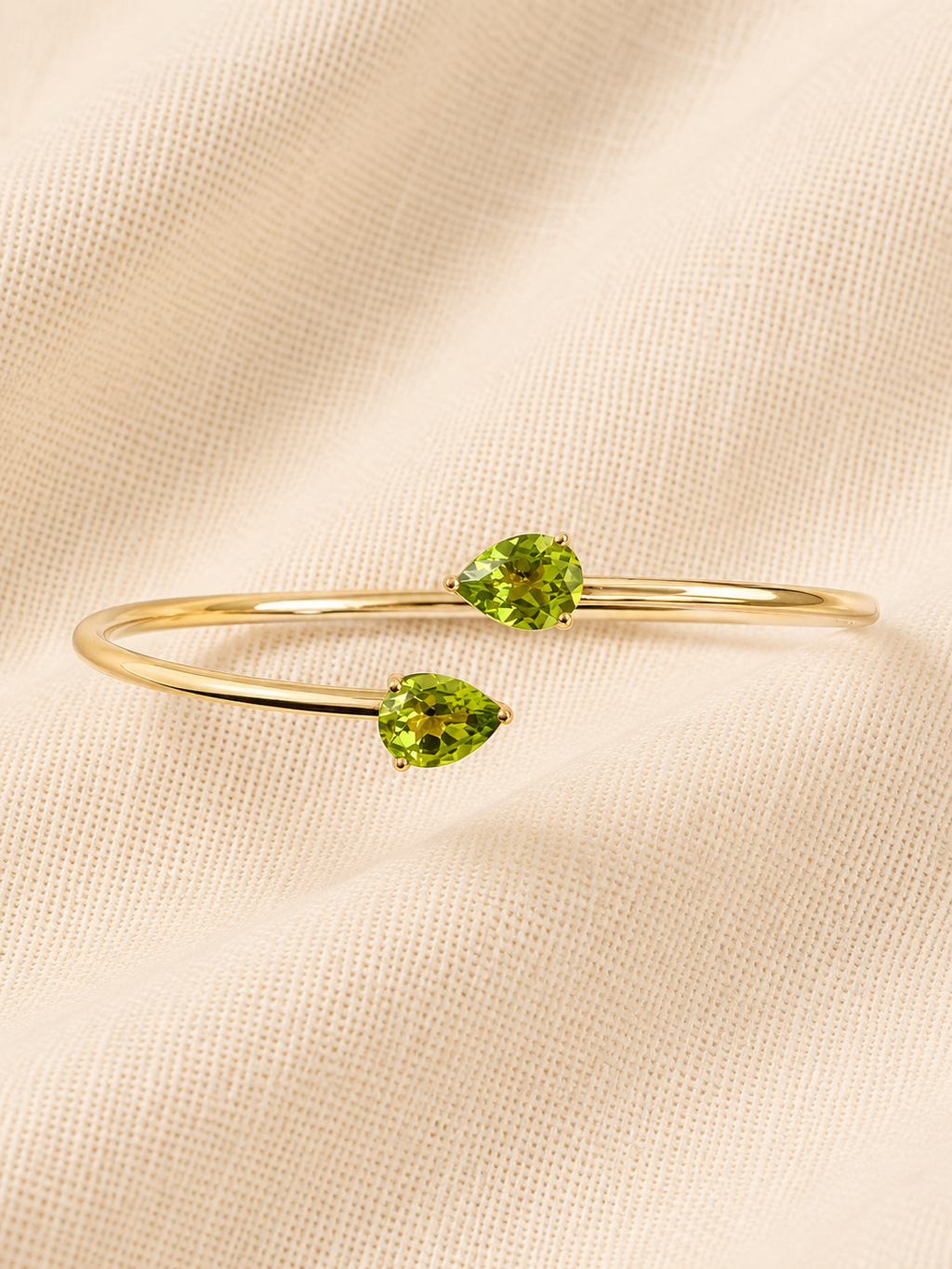 Top view of a solid 10k yellow gold bangle bracelet featuring two natural pear-cut green peridot gemstones by Ophelisse, displayed on a soft beige textured fabric background.
