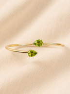 Top view of a solid 10k yellow gold bangle bracelet featuring two natural pear-cut green peridot gemstones by Ophelisse, displayed on a soft beige textured fabric background.