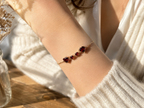 A delicate solid 10k rose gold chain bracelet by Ophelisse with two heart-shaped and two round-shaped natural red garnet gemstones worn on a person's wrist, styled with a white knit sweater in warm sunlight.