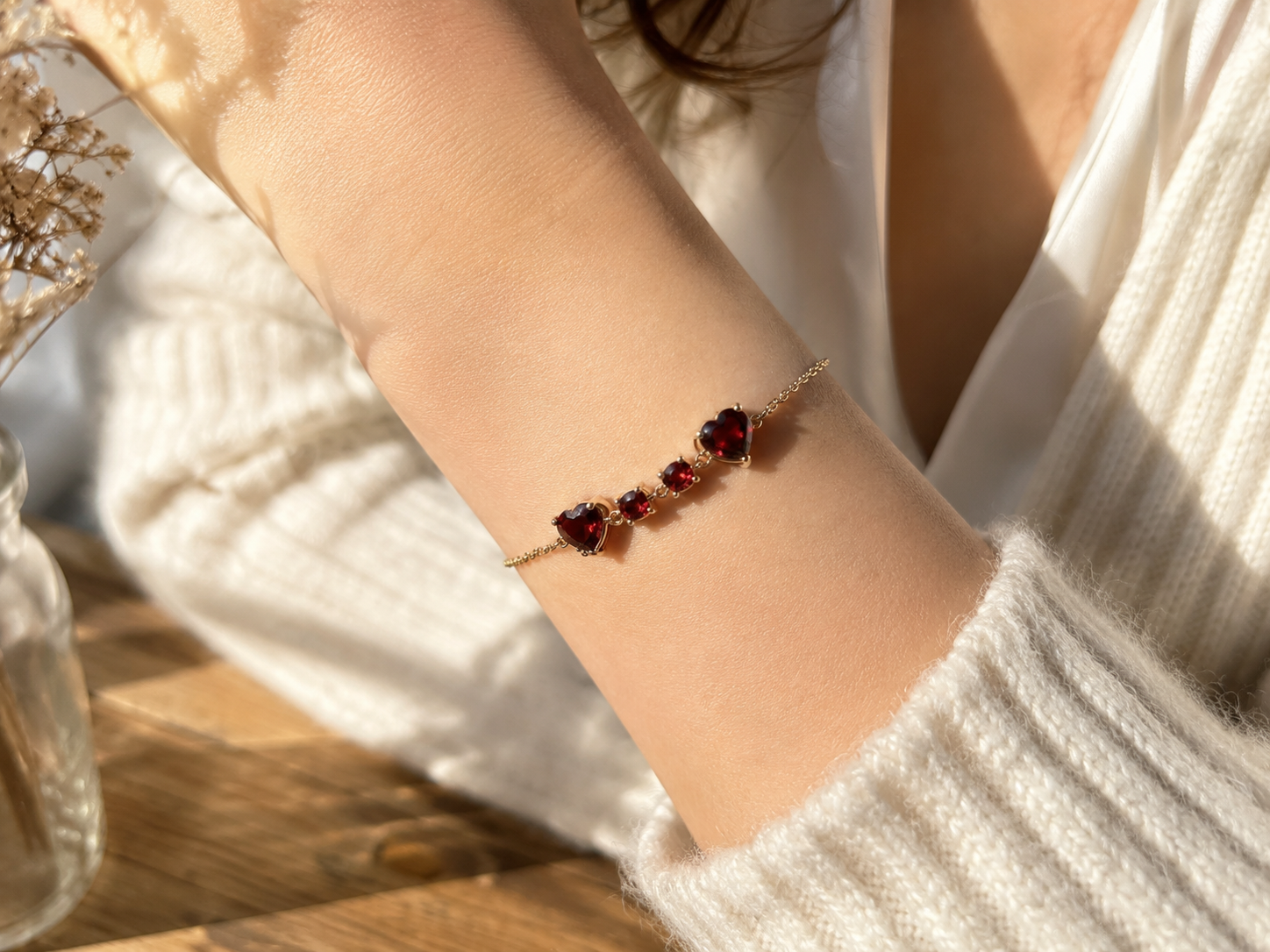 A delicate solid 10k rose gold chain bracelet by Ophelisse with two heart-shaped and two round-shaped natural red garnet gemstones worn on a person's wrist, styled with a white knit sweater in warm sunlight.