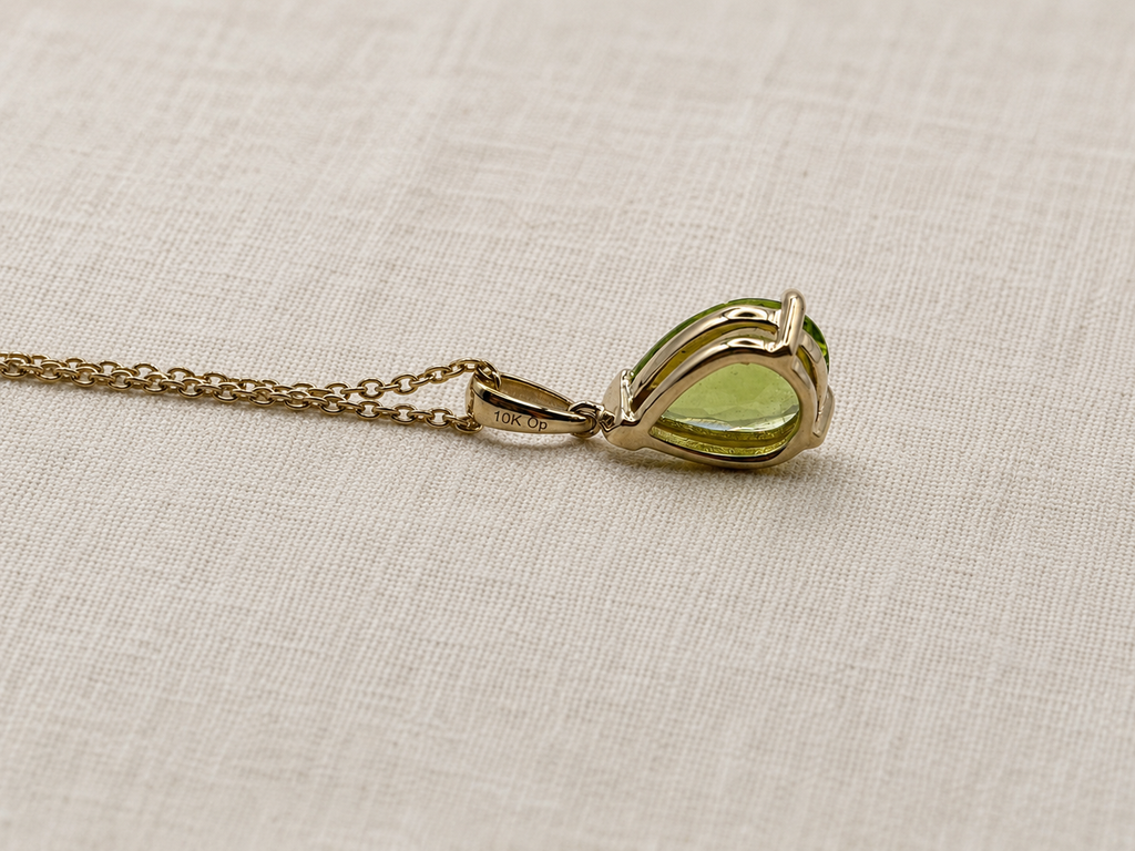 A 10k solid yellow gold pendant necklace with a teardrop-shaped natural green peridot gemstone by Ophelisse, showing the back of the pendant and stamped gold and trademark, resting on a textured beige linen fabric background.
