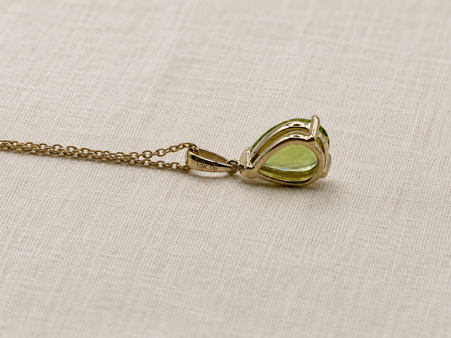 A 10k solid yellow gold pendant necklace with a teardrop-shaped natural green peridot gemstone by Ophelisse, showing the back of the pendant and stamped gold and trademark, resting on a textured beige linen fabric background.