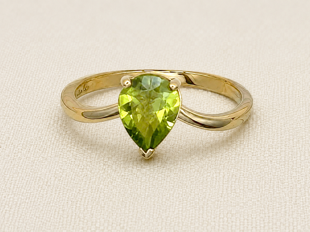 A solid 10k yellow gold ring featuring a pear-cut natural green peridot gemstone in a prong setting, displayed on a textured beige fabric background.