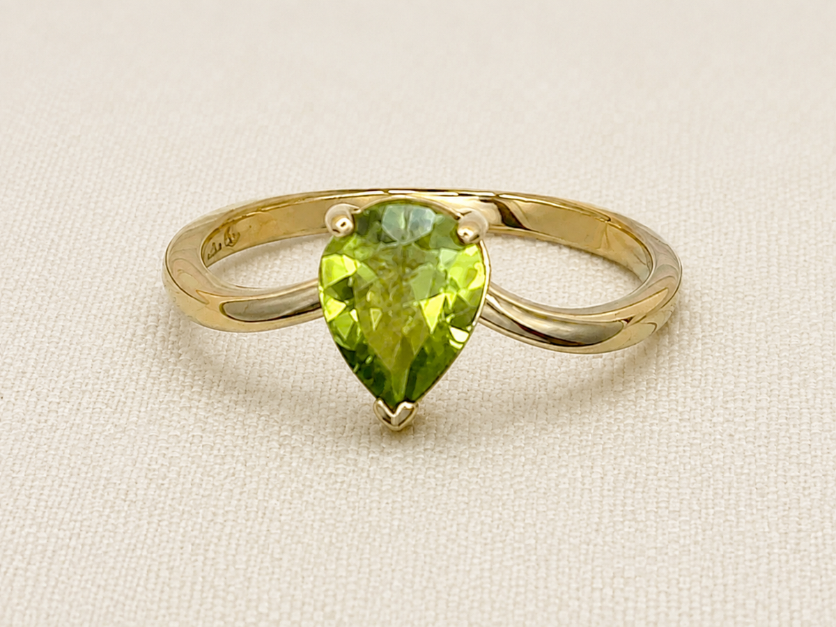A solid 10k yellow gold ring featuring a pear-cut natural green peridot gemstone in a prong setting, displayed on a textured beige fabric background.