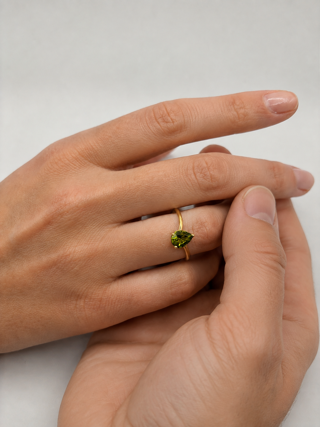 A close-up of a person's hands resting together, wearing a delicate solid 10k gold ring with a small green marquise-cut natural peridot gemstone by Ophelisse on the ring finger, set against an elegant light gray background. This ring is used to emphasize the promise between bonds and love.
