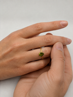 A close-up of a person's hands resting together, wearing a delicate solid 10k gold ring with a small green marquise-cut natural peridot gemstone by Ophelisse on the ring finger, set against an elegant light gray background. This ring is used to emphasize the promise between bonds and love.