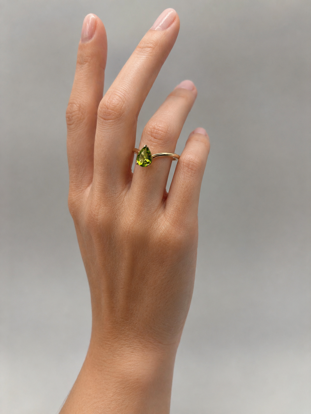 An Ophelisse solid 10k yellow gold ring with a pear-cut natural green peridot gemstone worn on a person's ring finger, shown against a gray neutral background. 
