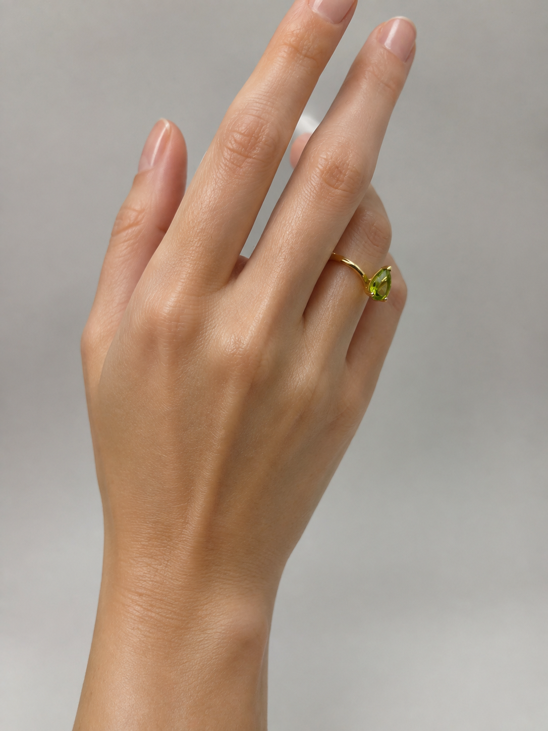 A close-up of a hand wearing a solid 10k yellow gold ring by Ophelisse with a natural green peridot gemstone, set against a soft gray background.