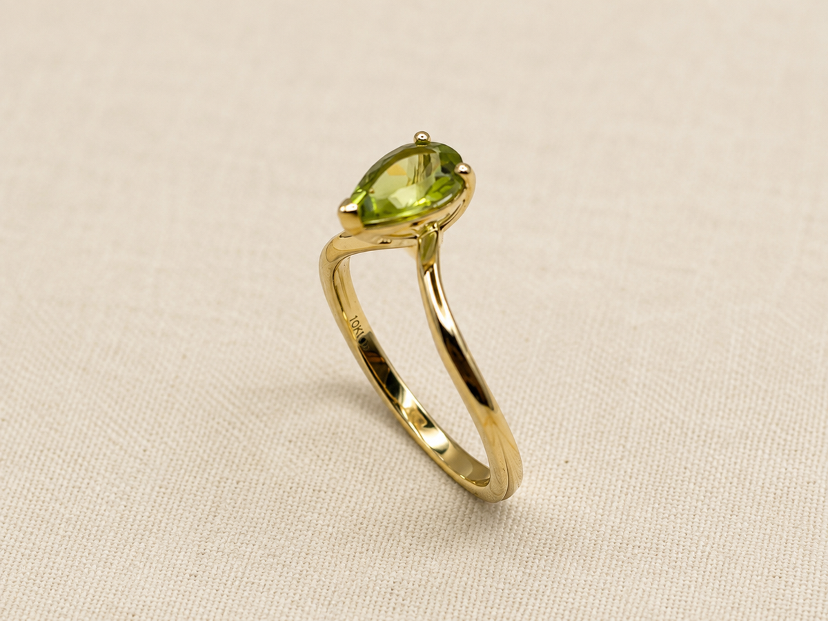 A solid yellow gold ring featuring a pear-cut green peridot gemstone by Ophelisse, displayed on an elegant textured beige fabric background.