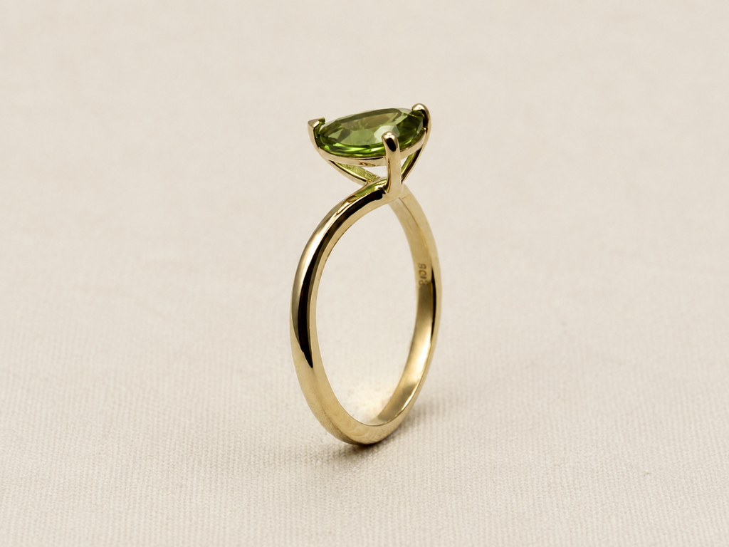 A 10k yellow gold ring by Ophelisse featuring a marquise-cut green gemstone in a prong setting, displayed on an elegant textured off-white background.