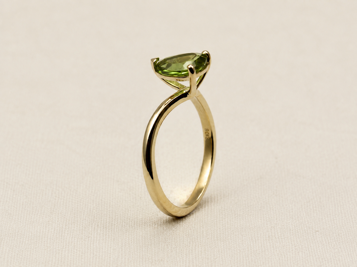 A 10k yellow gold ring by Ophelisse featuring a marquise-cut green gemstone in a prong setting, displayed on an elegant textured off-white background.