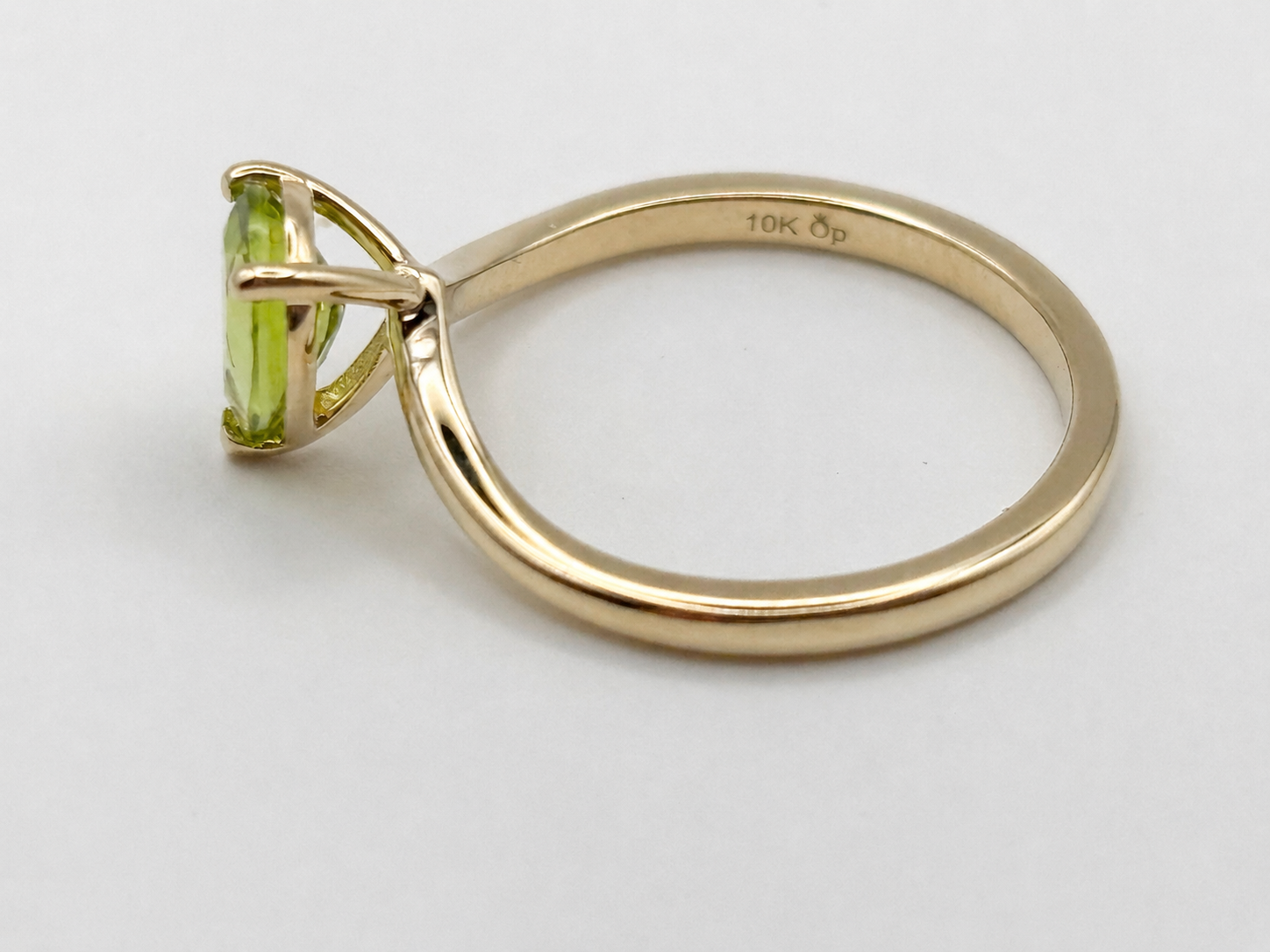 A solid yellow gold ring with a pear-cut natural peridot gemstone by Ophelisse, shown from a side profile on a light gray background. The inner band is stamped with '10K OP'.