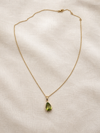 A solid 10k yellow gold chain necklace with a teardrop-shaped natural green peridot gemstone pendant by Ophelisse, laid flat on a textured beige linen fabric background.