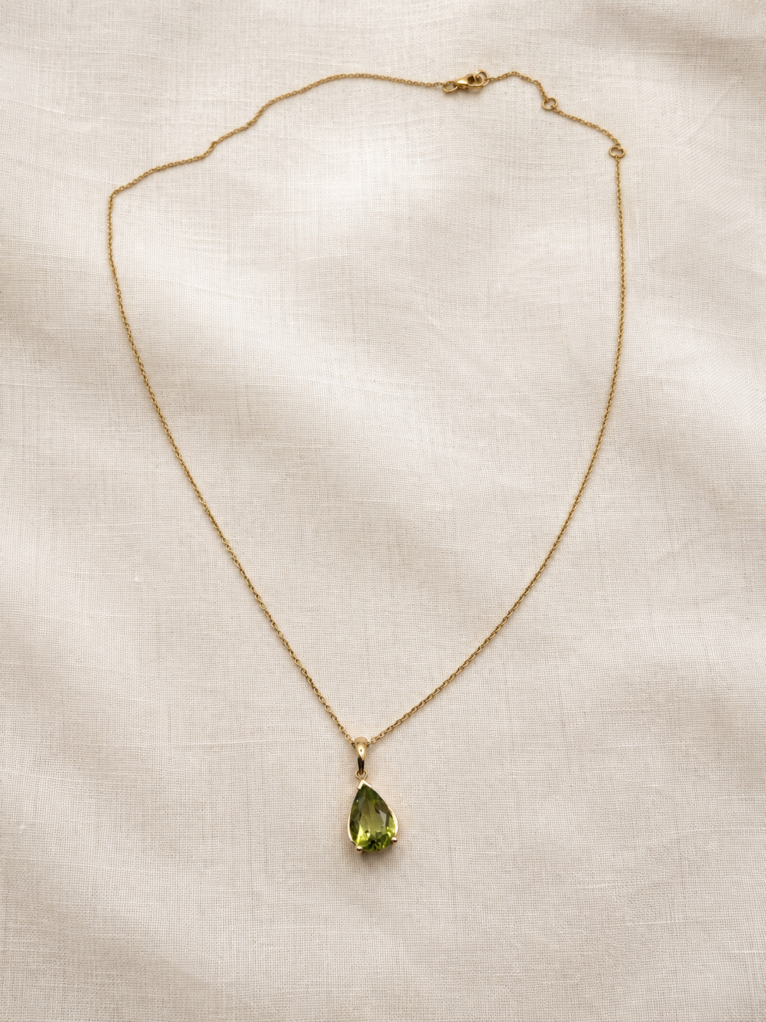 A solid 10k yellow gold chain necklace with a teardrop-shaped natural green peridot gemstone pendant by Ophelisse, laid flat on a textured beige linen fabric background.