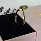 A solid yellow gold 10k ring with a pear-cut natural green peridot gemstone displayed in a black ring box, with a pink Ophelisse branded box and lavender sprigs in the background.