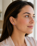 A close-up side profile of a woman with dark brown hair wearing a small purple natural amethyst geometric stud earring by Ophelisse, set against a soft, bright, neutral background.